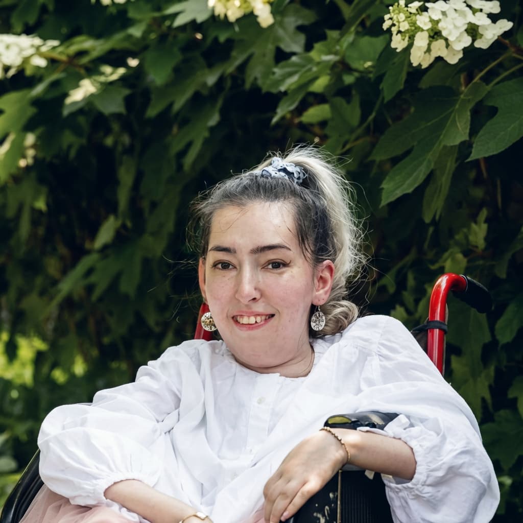Atieh Taheri, a young woman with light skin and dark blonde hair pulled back in a ponytail with a blue scrunchie, smiles warmly at the camera. She is seated in a wheelchair with red handles, wearing a white blouse and large round earrings. Behind her is a lush green hedge with clusters of white hydrangea blossoms.