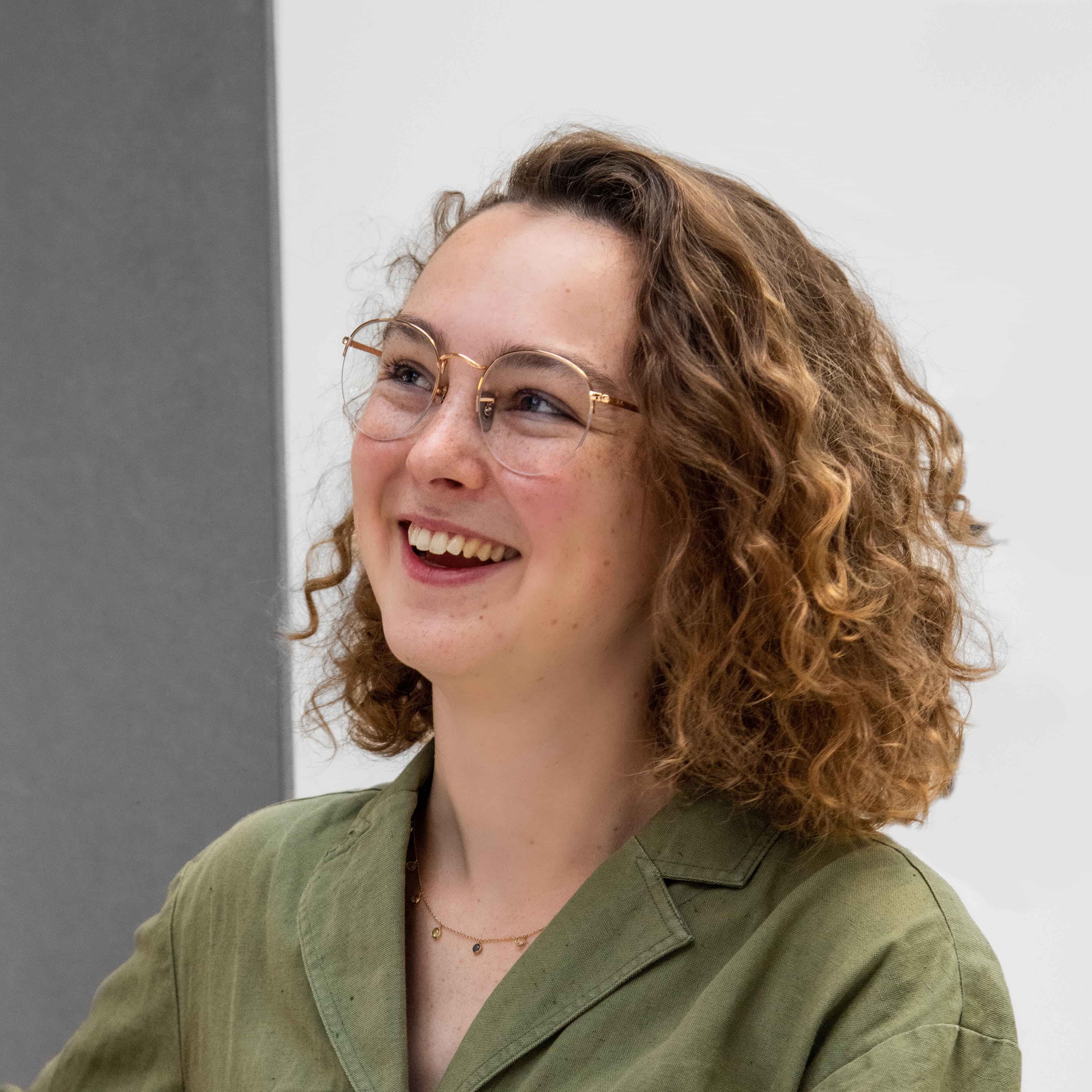 Eva Peet, a woman with light skin, shoulder-length curly hair and glasses, smiles and looks to the side.