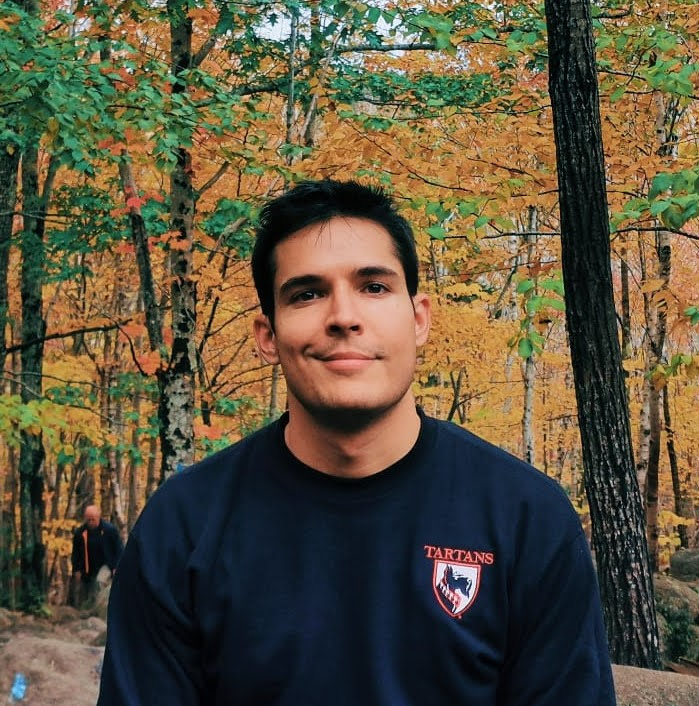 A young man with dark hair smiling at the camera, wearing a navy sweatshirt, standing in front of autumn foliage.