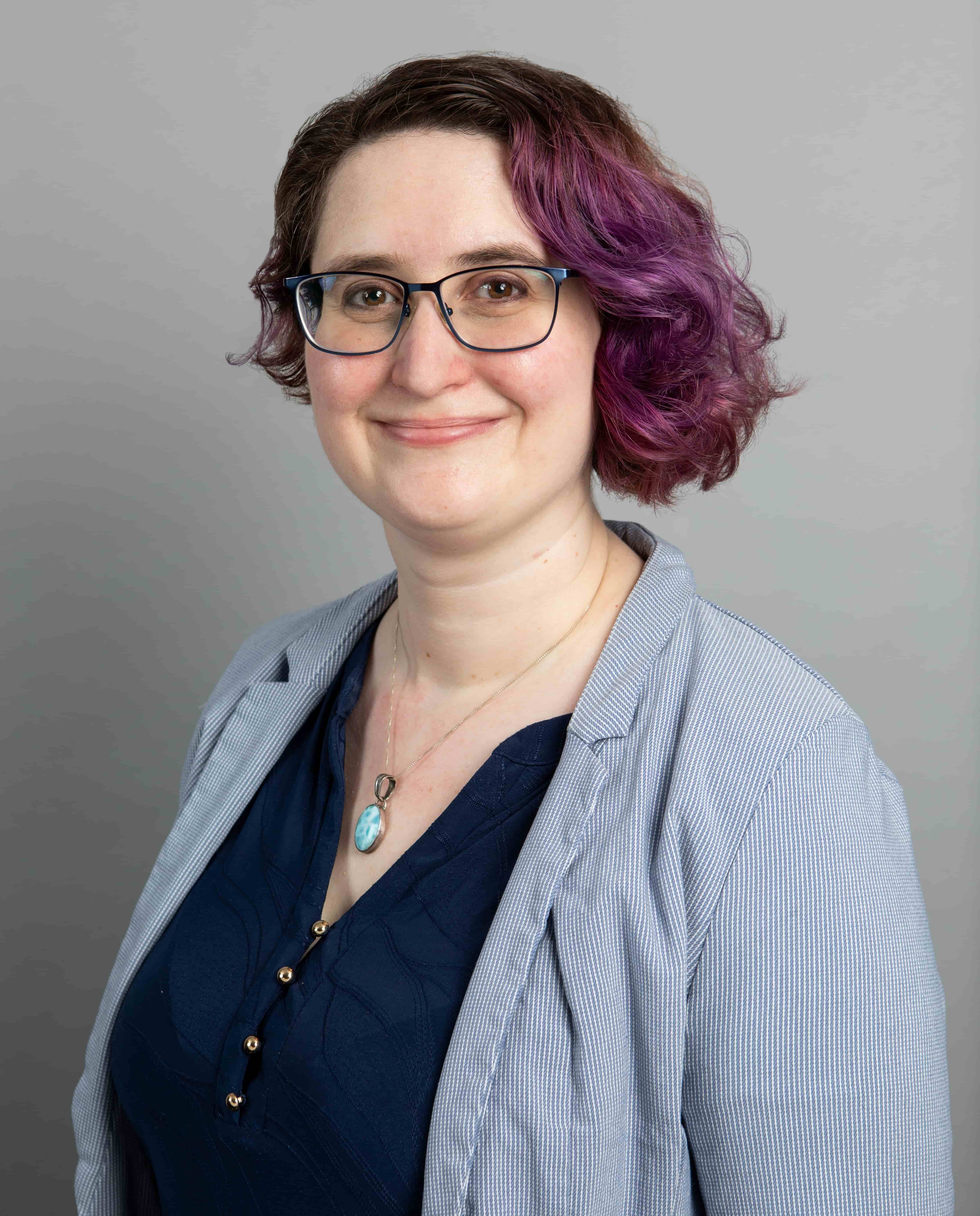 Dr. Martin has asymmetrical brown hair with purple on the longer side. They have blue glasses and are wearing a navy blue top, light blue striped jacket, and a larimar necklace.