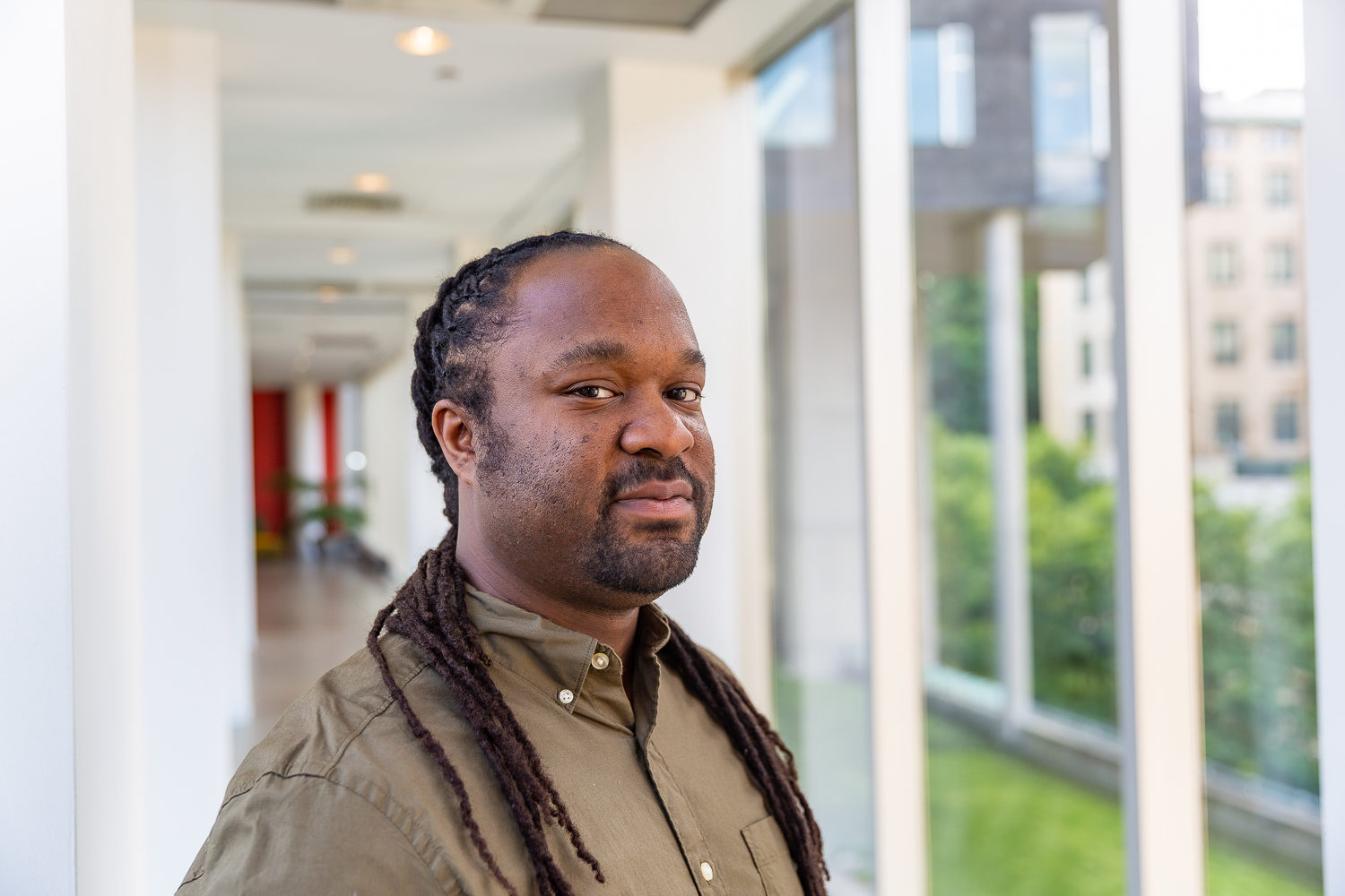 A man with dark skin, long locs, and a short beard, wearing an olive shirt, looking at the camera in a bright hallway.
