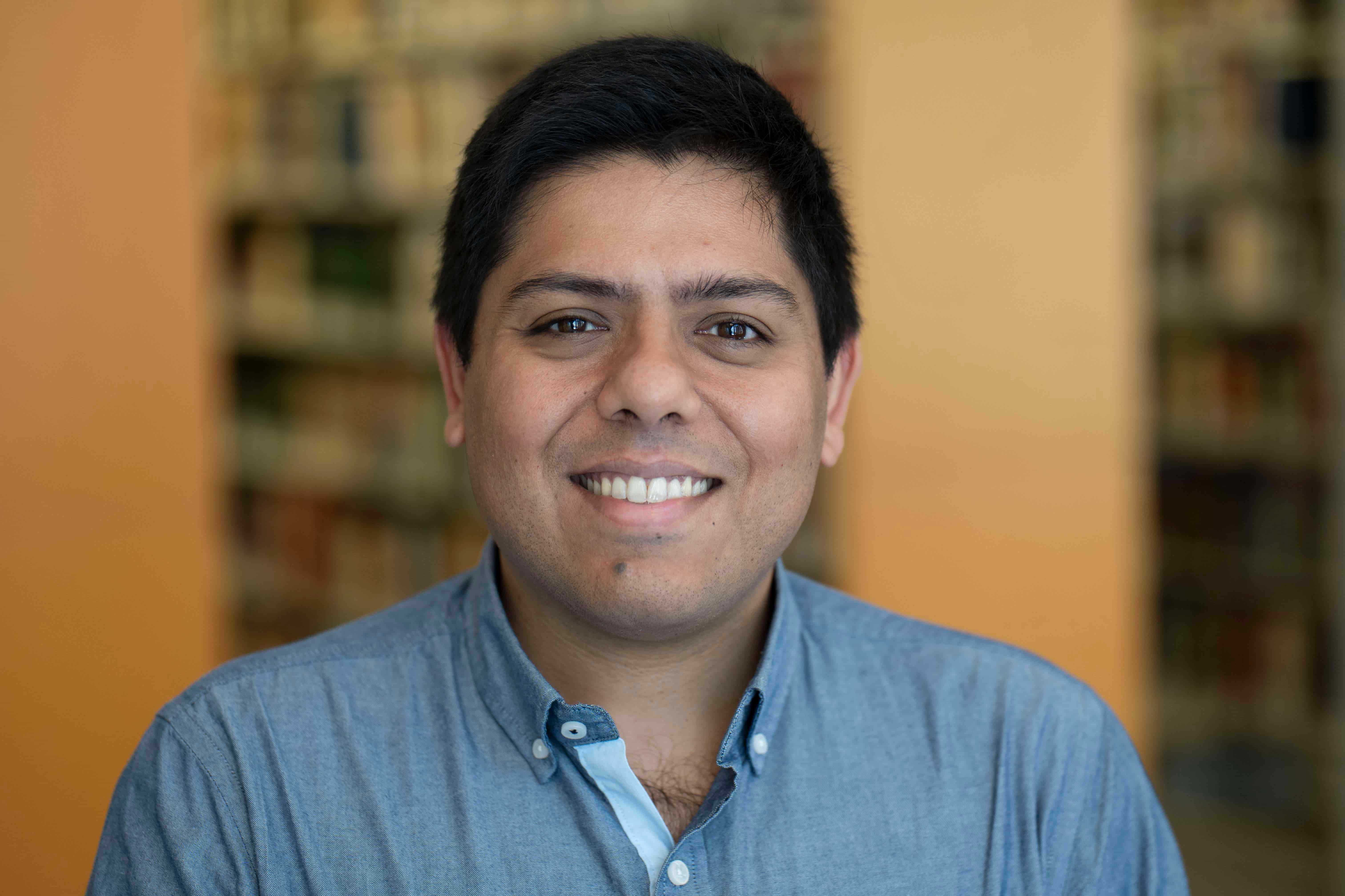Headshot of Raj Korpan smiling at the camera, with short dark hair and wearing a blue button-down shirt, against a softly blurred library background.