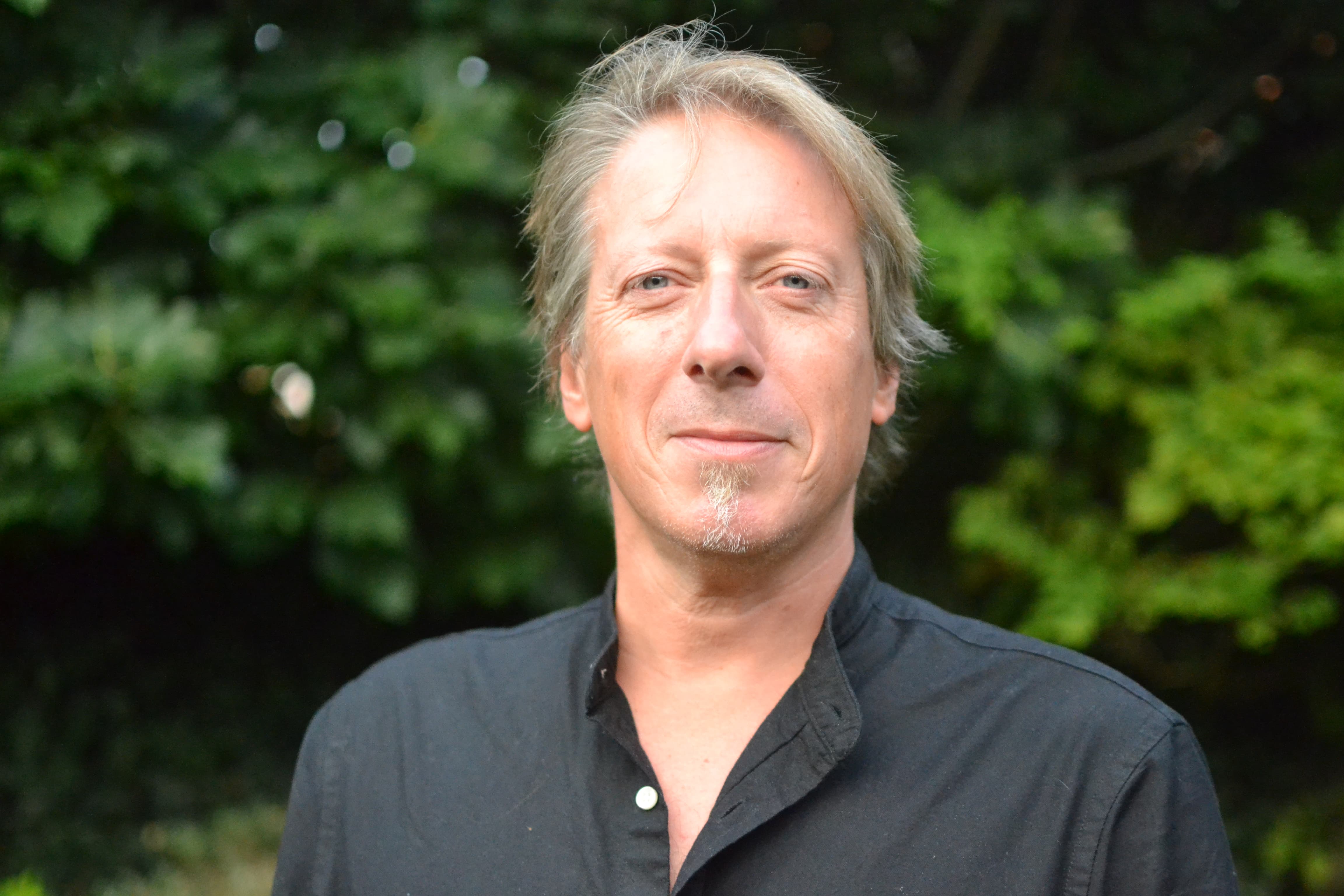 Light-haired man looking at the camera in front of green shrubbery